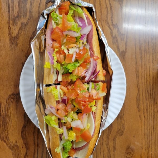 A large NYC Village Pizza sub sandwich, cut in half, wrapped in foil on a paper plate.