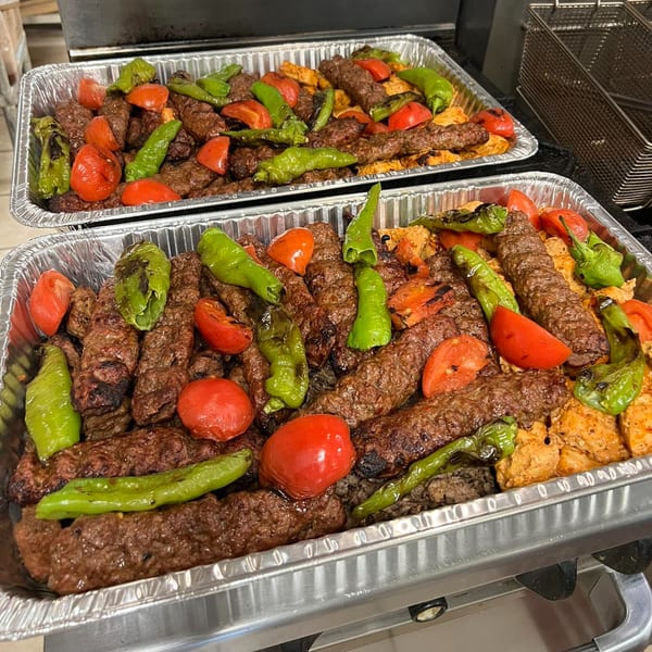 Two foil trays filled with grilled meat kebabs, green peppers, and tomatoes.