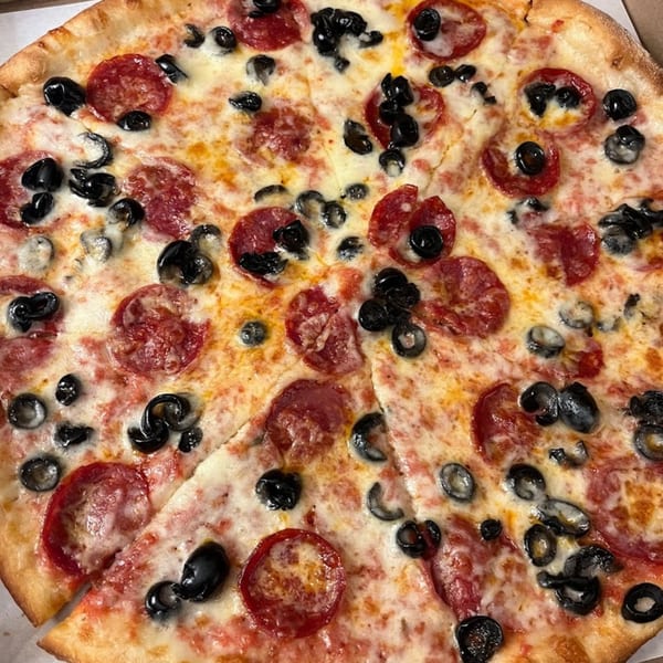 A whole NYC Village Pizza with pepperoni and black olives, with one slice removed.