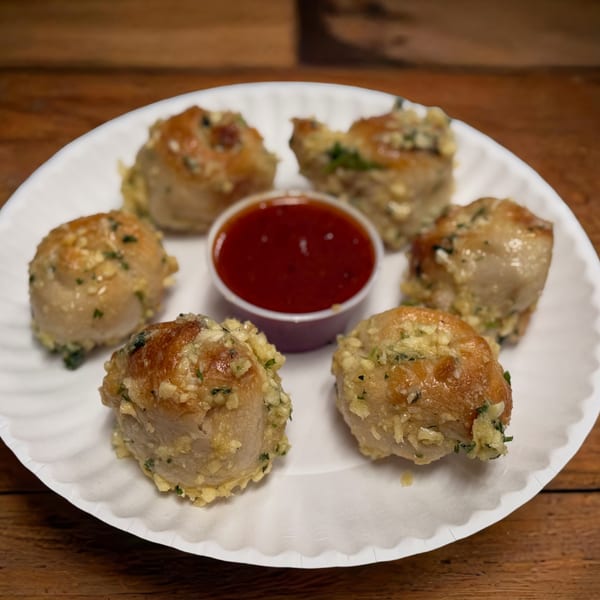 Six golden-brown garlic knots with herbs and cheese, arranged around a small cup of red sauce.